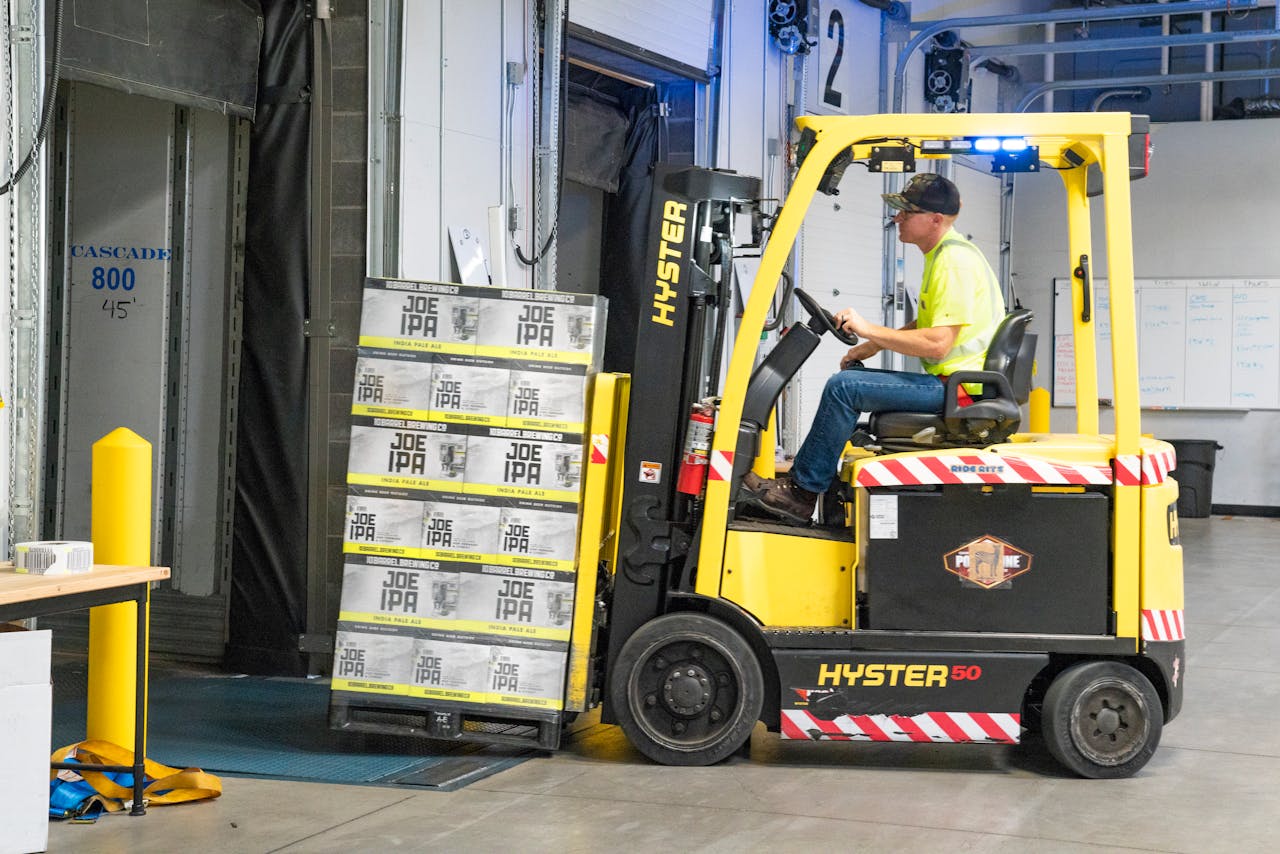hero-img-02 A worker drives a Hyster forklift moving Joe IPA boxes in a warehouse.